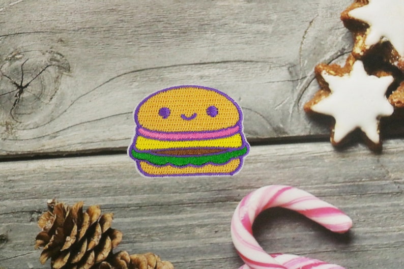 May include: A cute embroidered patch of a smiling hamburger with purple, yellow, pink, green, and brown details.