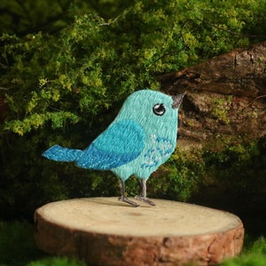 Puede incluir: Un pájaro azul bordado con detalles blancos se posa sobre una rodaja de madera. El pájaro está posado sobre una superficie verde cubierta de musgo con setas rojas en el fondo.