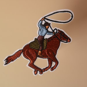 May include: Embroidered patch of a cowboy riding a brown horse and holding a lasso. The cowboy is wearing a blue shirt and brown pants. The patch is white with brown, blue, and green stitching.