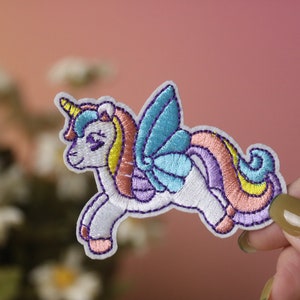 Puede incluir: Un unicornio blanco bordado con una crin y cola arcoíris, alas azules y un cuerno morado. El unicornio está volando y tiene un estilo caprichoso y de dibujos animados.