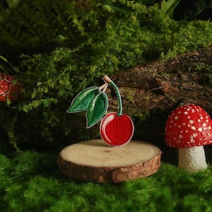 Może przedstawiać: Czerwona wiśnia z zielonymi liśćmi wyszyta na drewnianym dysku, otoczona trzema czerwonymi i białymi grzybami i zielonym mchem.