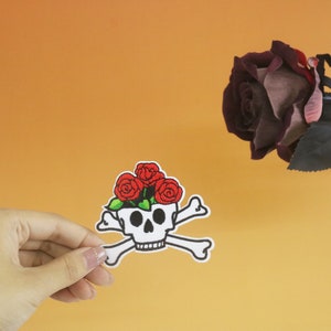 Może przedstawiać: Biała czaszka z przekreślonymi kośćmi i czerwonymi różami na górze. Czaszka znajduje się na białym tle.