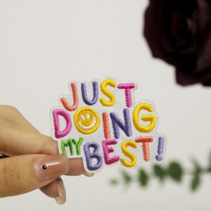 May include: A white iron-on patch with colorful embroidered text that reads "JUST DOING MY BEST!" with a yellow smiley face in the middle of the word "DOING".