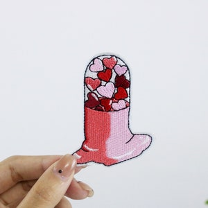 Op de afbeelding: Een geborduurde patch in de vorm van een pilcapsule gevuld met kleine rode en roze hartjes, in roze en rood.
