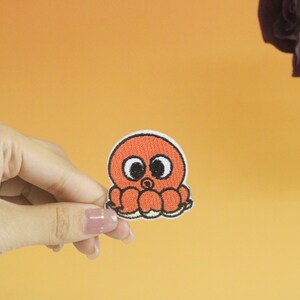 May include: A small orange octopus with black eyes and a white outline. The octopus is embroidered on a white fabric patch.