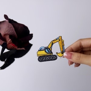 Pu&ograve; includere: Un toppa ricamata gialla di un escavatore con una cabina blu e cingoli neri.