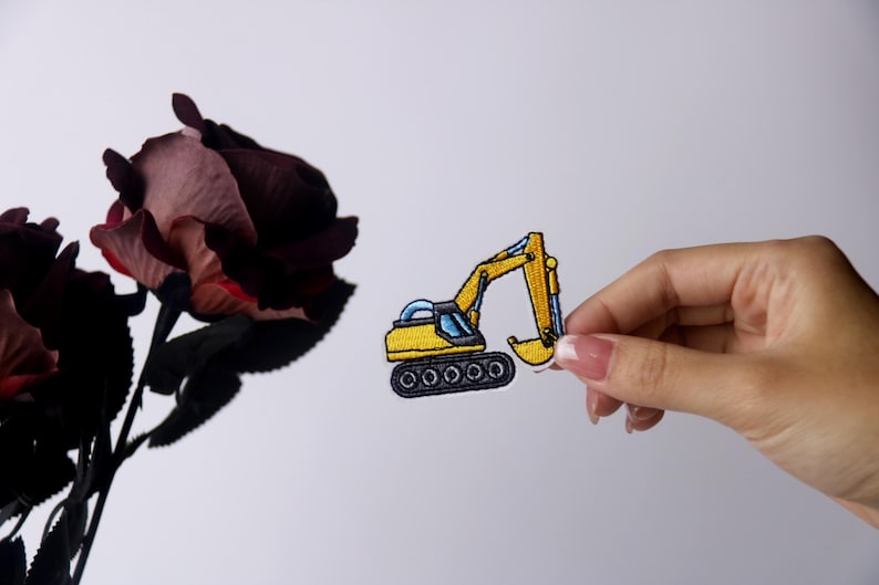 Pu&ograve; includere: Un toppa ricamata gialla di un escavatore con una cabina blu e cingoli neri.