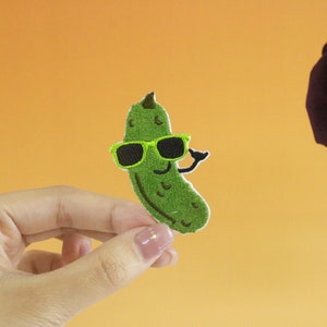 May include: A green embroidered pickle wearing sunglasses and smiling. The pickle is being held by a hand against a light orange background.