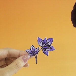 May include: A purple embroidered flower patch with two blossoms and a stem. The patch is being held by a hand against a yellow background.