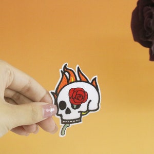 Op de afbeelding: Geborduurde patch met een witte schedel met een rode roos in de oogkas en oranje vlammen. De patch wordt tegen een oranje achtergrond gehouden, met een donkere roos in de rechterbovenhoek.