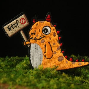 May include: Embroidered patch of an orange dinosaur with red accents, holding a sign that reads "stop". The dinosaur has a white belly and is set against green moss and a dark background. A fun, colourful patch.