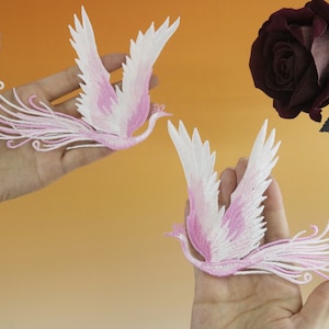 May include: Two embroidered bird patches with pink and white details are held up against an orange background. The birds have outstretched wings and flowing tail feathers. A dark red rose is in the background.