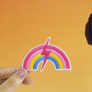 以下が含まれることがあります： ピンク、イエロー、ブルーのデザインの虹色の刺繍パッチ。虹の中央に白い稲妻が縫い付けられています。