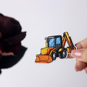 Könnte beinhalten: Ein gestickter Bügelaufkleber mit einer kleinen gelben und schwarzen Baggerschaufel mit blauer Windschutzscheibe und orangefarbenem Löffel.