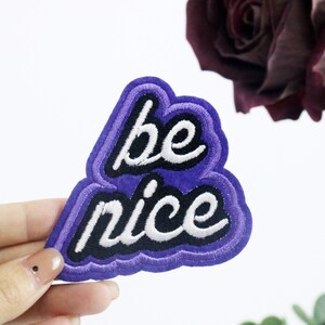 Peut inclure: Patch en feutre violet avec du texte brodé blanc qui dit "be nice" avec des contours noirs.