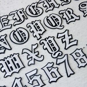 May include: White iron-on letters and numbers in a gothic font style. The letters are outlined in black and have a white fill. The letters include A, B, C, D, E, F, G, H, I, J, K, L, M, N, O, P, Q, R, S, T, U, V, W, X, Y, Z, 1, 2, 3, 4, 5, 6, 7, 8, 9.