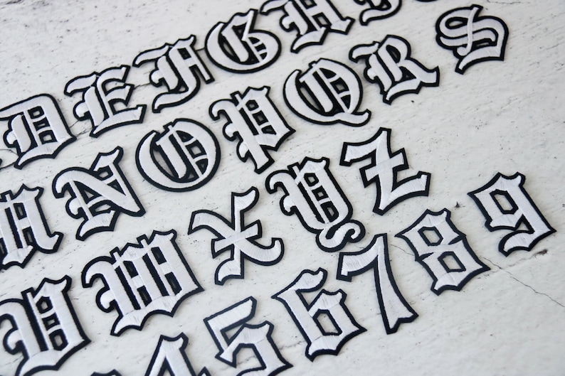 May include: White iron-on letters and numbers in a gothic font style. The letters are outlined in black and have a white fill. The letters include A, B, C, D, E, F, G, H, I, J, K, L, M, N, O, P, Q, R, S, T, U, V, W, X, Y, Z, 1, 2, 3, 4, 5, 6, 7, 8, 9.