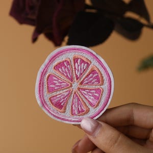 Puede incluir: Un parche de hierro bordado de color rosa y blanco con forma de rodaja de pomelo.