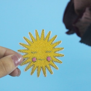 Peut inclure: Un soleil brodé jaune avec un visage souriant et des joues roses. Le soleil a une finition pailletée dorée et est entouré de rayons blancs.