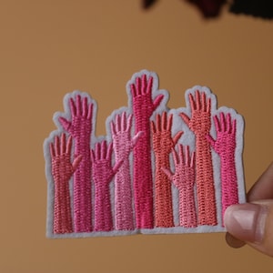 May include: A patch with a group of pink and red embroidered hands raised in the air. The hands are all different shades of pink and red, and they are arranged in a cluster.