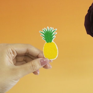 May include: A hand holding a yellow and green felt pineapple patch. The pineapple is on a white background.