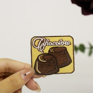 May include: A yellow embroidered patch with the word "Chocolate" in white cursive lettering and two brown chocolate candies.