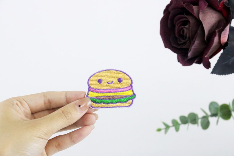 May include: A yellow, purple, and green embroidered iron-on patch of a smiling cartoon hamburger.
