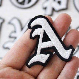 May include: White embroidered letter A on a black felt background. The letter is in a gothic style font.