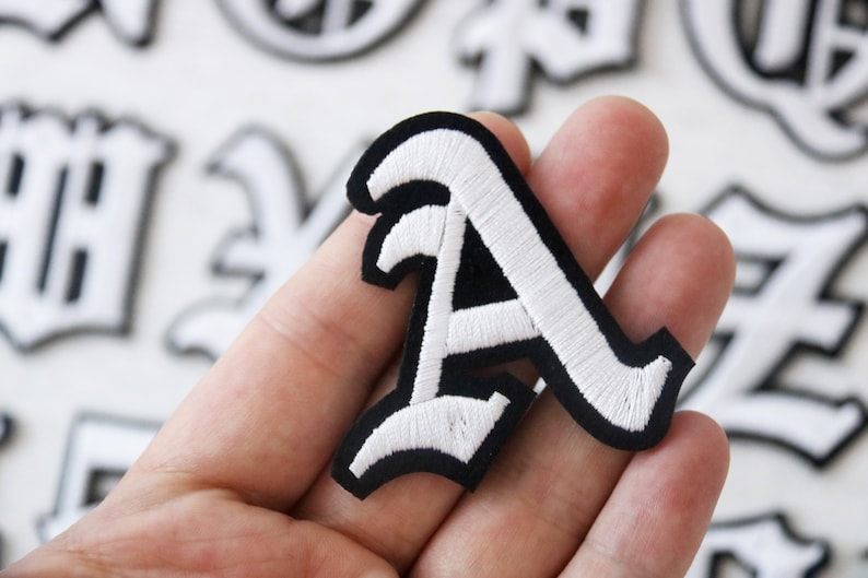 May include: White embroidered letter A on a black felt background. The letter is in a gothic style font.