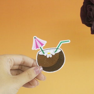 May include: A brown coconut with a pink umbrella and a green straw, a white flower is inside the coconut.