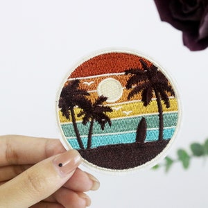 May include: Round embroidered patch depicting a sunset beach scene with palm trees, a surfboard, and seagulls. The design features orange, yellow, teal, and brown hues, with a white border. The patch is circular.