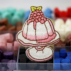 May include: A white and pink embroidered patch of a dessert with a raspberry on top. The dessert is on a white pedestal with a brown outline. The patch is in a clear plastic container with other colorful patches.