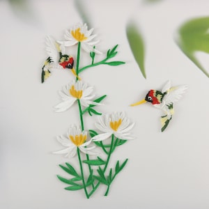 Puede incluir: Parche bordado con un grupo de flores blancas con centros amarillos y dos colibríes verdes con detalles rojos, negros y amarillos.