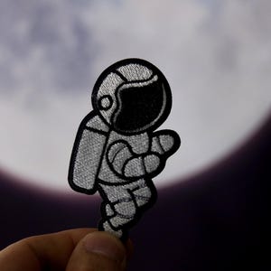May include: A silver and black embroidered astronaut patch with a white background. The astronaut is wearing a helmet and is in a walking position.