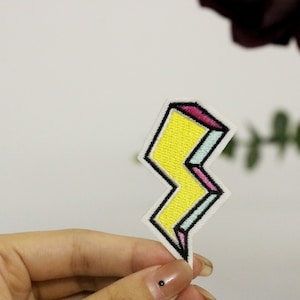 Könnte beinhalten: Ein gelber Blitz-förmiger Bügelaufkleber mit rosa, blauen und weißen Akzenten.