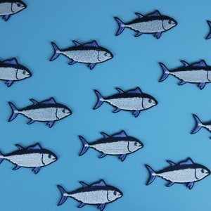 May include: A blue background with multiple blue and white embroidered tuna fish patches. The patches are arranged in a pattern, with some facing left and some facing right.