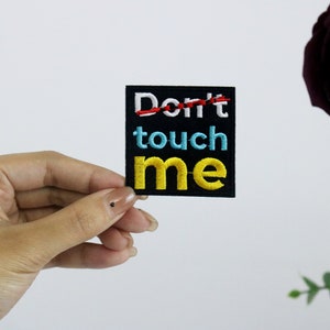 Könnte beinhalten: Ein schwarzer gestickter Aufnäher mit dem Text "Don't touch me" in bunten Buchstaben. Das Wort "Don't" ist mit einem roten Strich durchgestrichen.