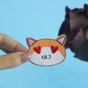 Puede incluir: Un parche blanco bordado con una cara de gato naranja de dibujos animados con corazones rojos para los ojos y una pequeña sonrisa. El parche se sostiene en una mano contra un fondo azul claro.