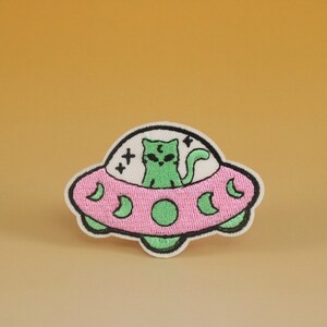 May include: An embroidered patch featuring a green cat riding in a pink flying saucer. The saucer has a black outline and five crescent moons around the rim. The cat has black eyes and a black lightning bolt on its head.