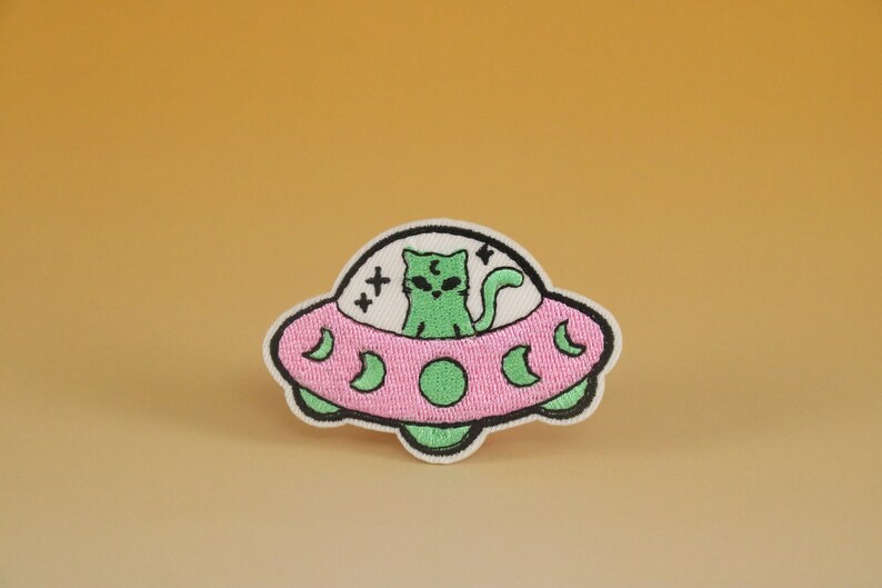 May include: An embroidered patch featuring a green cat riding in a pink flying saucer. The saucer has a black outline and five crescent moons around the rim. The cat has black eyes and a black lightning bolt on its head.