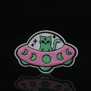 May include: Green cat alien in a pink and white flying saucer with stars and moons. The cat has green eyes and a white face. The saucer is outlined in black.