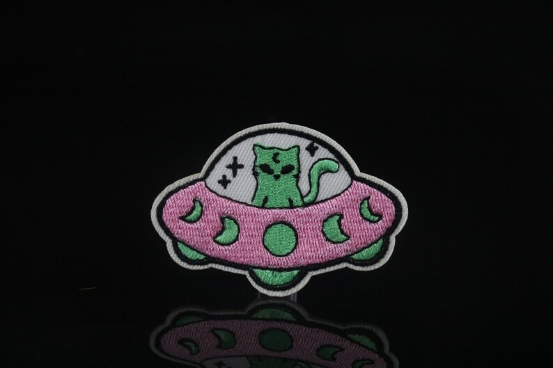 May include: Green cat alien in a pink and white flying saucer with stars and moons. The cat has green eyes and a white face. The saucer is outlined in black.