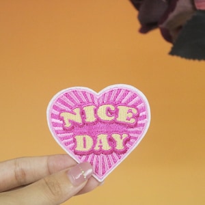 May include: A pink heart-shaped embroidered patch with the words "NICE DAY" in yellow, outlined in white. The patch has a radiating sunburst design and is held against an orange background.