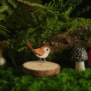 Pode incluir: Um pequeno pássaro bordado marrom e branco empoleirado em um tronco de madeira cercado por musgo verde e três cogumelos. Um cogumelo é vermelho com manchas brancas, outro é preto com manchas brancas e o último é vermelho com manchas brancas.