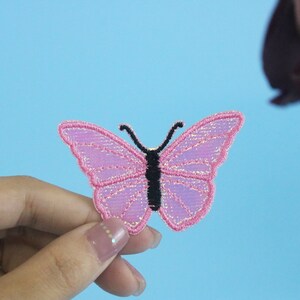 May include: A pink butterfly-shaped fabric patch with a black outline. The patch is held in a hand against a light blue background.