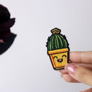Könnte beinhalten: Ein gestickter Kaktus-Patch in Grün und Gelb mit einem lächelnden Gesicht. Der Kaktus befindet sich in einem gelben Topf mit braunem Rand.