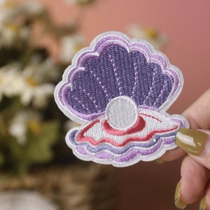 Op de afbeelding: Een geborduurde patch met een schelp met een parel erin, in tinten paars en wit. Deze patch is perfect om kleding, tassen of andere accessoires een vleugje whimsy te geven.