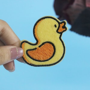 May include: A yellow felt rubber ducky patch with an orange beak and black outline. The patch is being held in a hand.