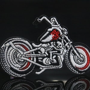 Könnte beinhalten: Weiß gestickter Motorrad-Patch mit roten Akzenten. Das Motorrad ist in Seitenansicht dargestellt, wobei das Vorderrad leicht nach rechts gedreht ist.