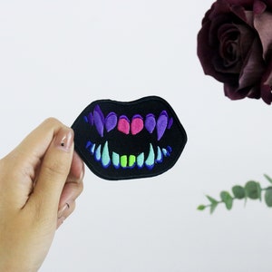 May include: A black embroidered patch featuring a colourful mouth with sharp teeth. The teeth are green, blue, purple and pink. The patch is being held in a hand.